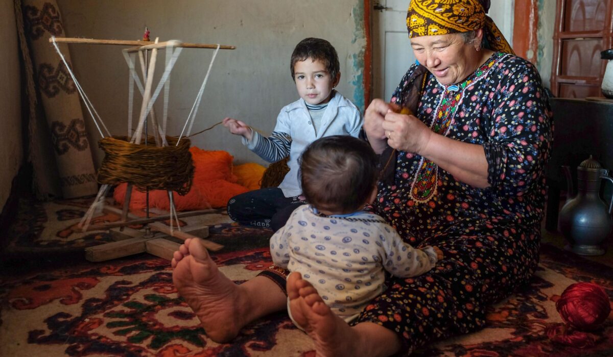 In the Gara Gum desert, Turkmenistan, a grandmother looks after her youngest grandson. In the background, her granddaughter helps spool the yarn, which will then be used to make plaited lengths (alaja) that protect against the evil eye. These alaja are most commonly plaited with four colors,  the brown color often made from un-dyed camel hair.