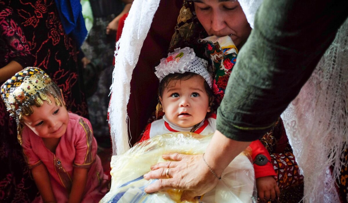 In the Gara gum desert of Turkmenistan, a bride is given a neighbor's child to hold on her wedding day in the hopes that the bride too, will soon have children of her own. The mother's hand rests on a parcel of wedding gifts (podarka) and to the side, a young girl looks on excitedly. The bride is looking down and holding a white scarf (ak ýaglyk) between her lips in recognition of her modesty. She is covered from head to toe in layers of intricately embroidered silks and cotton (kürte), with her hair bound in scarves and adorned with metal jewelry which falls to her knees.