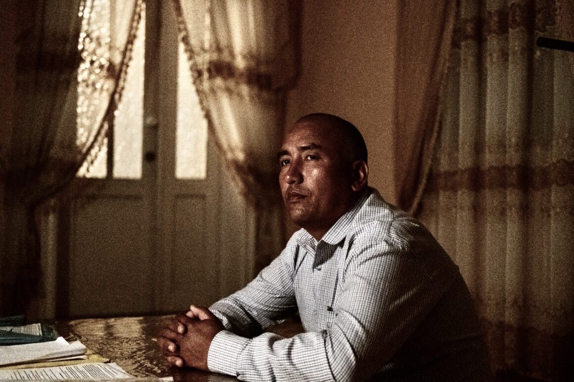 A man (former political prisoner) sitting at the table, hands clasped on the table, looking up and to the left.